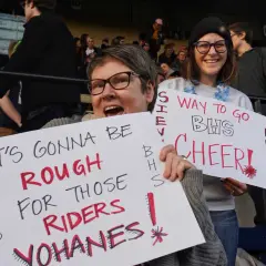 Fans hold signs at a snap raise fundraiser. One sign reads "IT'S GONNA BE ROUGH FOR THOSE RIDERS YOHANES!