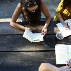 Two women study at a picnic table, one reading a book, the other writing notes. A board retreat activity.