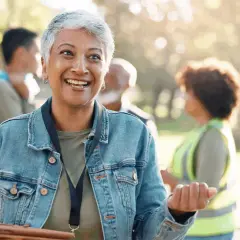 A woman with short gray hair smiles, her eyes bright. She's wearing a denim jacket and talking to others in a park, perhaps discussing why do you want to be a volunteer.
