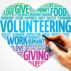 A hand holds a pen over a word cloud about volunteering, highlighting themes like "giving," "love," and "teamwork." This image represents the power of online volunteering.