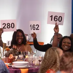 Excited guests participate in a paddle raise fundraiser, holding up signs with numbers like 280, 162, and 160.