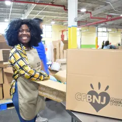 A smiling woman in an apron and gloves packs boxes at a food bank, embodying the spirit of a food bank volunteer job description.