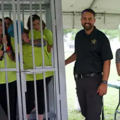 Smiles abound at a jail and bail fundraiser. A woman in a "Jail and Bail" shirt stands by a cage with women in neon shirts. A deputy and a woman holding a baby smile nearby.