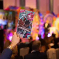 A hand holds up a colorful gala invitation for "School the World" at a vibrant event.