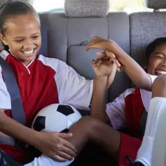 Two excited girls in soccer uniforms share a ball in the backseat, ready for their soccer fundraiser.