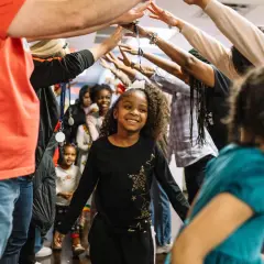 A group of children and adults form a tunnel with their arms raised, celebrating. This scene captures the joy of community and highlights places for kids to volunteer and make a difference.