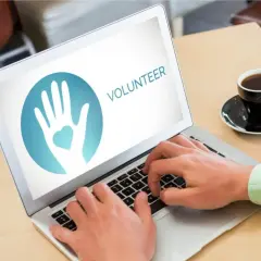 Hands typing on a laptop displaying a volunteer logo, ready to engage in online service projects. A coffee cup sits nearby.