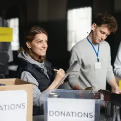 Volunteers organize donations for a free online community service. A woman smiles while checking items on a clipboard, while others sort canned goods.