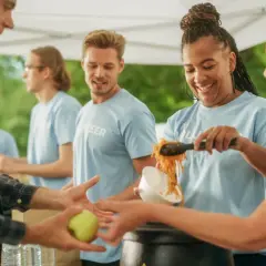 Volunteers serve food, showing what are some community services. A man receives an apple, while others get noodles.
