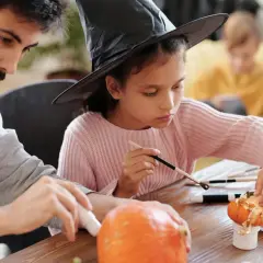 A girl in a witch hat and a man decorate pumpkins, showcasing fun Halloween fundraiser ideas.