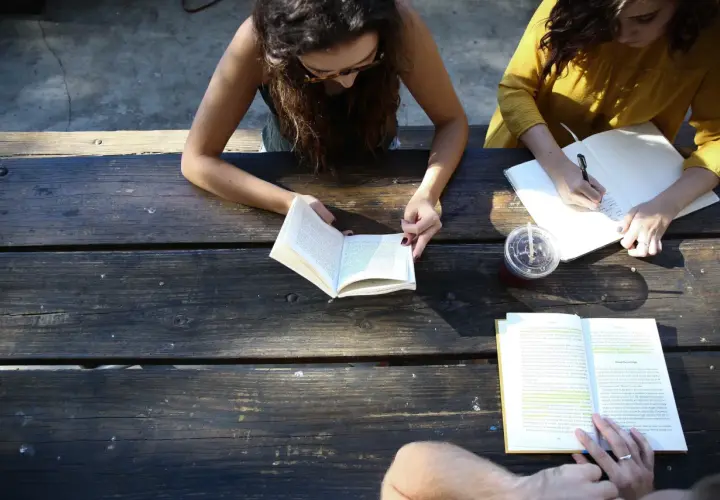 Two women study at a picnic table, one reading a book, the other writing notes. A board retreat activity.