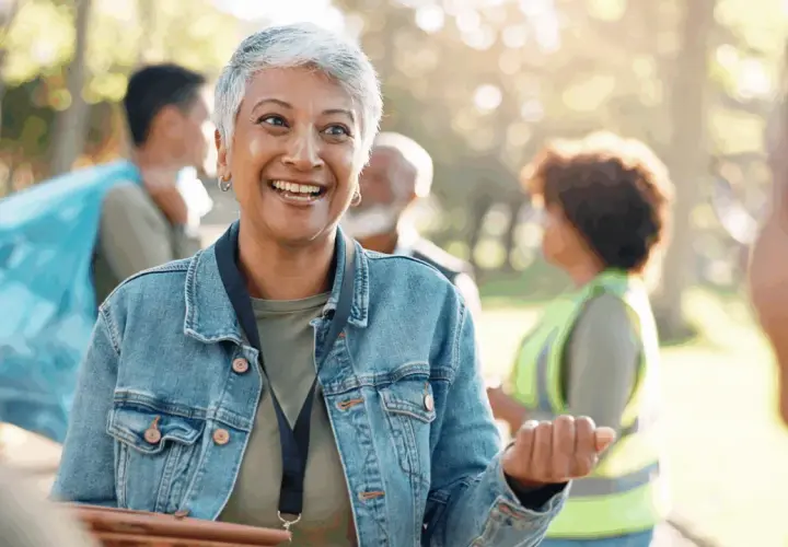 A woman with short gray hair smiles, her eyes bright. She's wearing a denim jacket and talking to others in a park, perhaps discussing why do you want to be a volunteer.