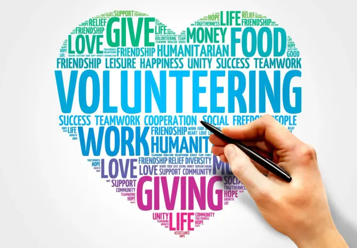 A hand holds a pen over a word cloud about volunteering, highlighting themes like "giving," "love," and "teamwork." This image represents the power of online volunteering.