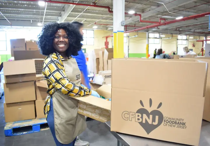 A smiling woman in an apron and gloves packs boxes at a food bank, embodying the spirit of a food bank volunteer job description.