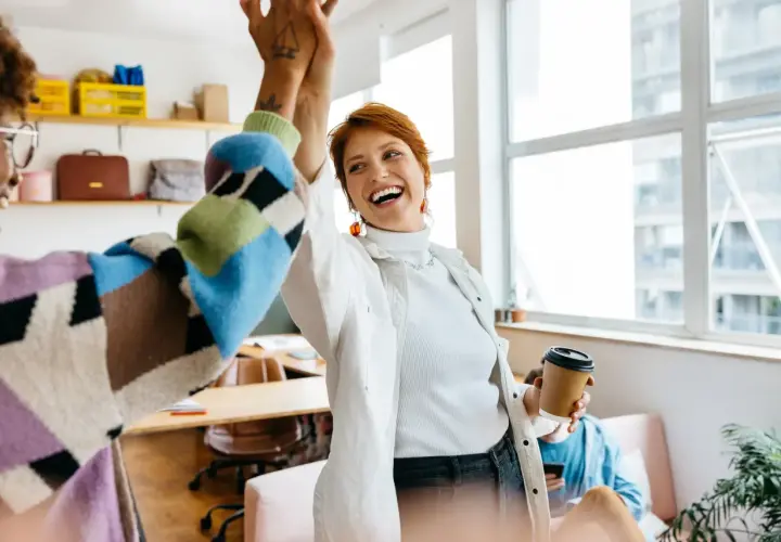 Two women high-five in an office, celebrating a successful project. One holds a coffee cup, perhaps after securing a moz nonprofit discount.