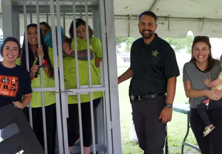 Smiles abound at a jail and bail fundraiser. A woman in a "Jail and Bail" shirt stands by a cage with women in neon shirts. A deputy and a woman holding a baby smile nearby.