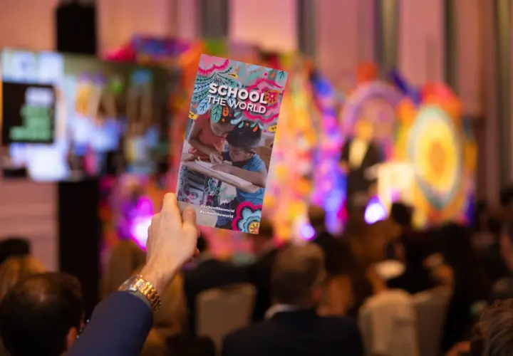 A hand holds up a colorful gala invitation for "School the World" at a vibrant event.