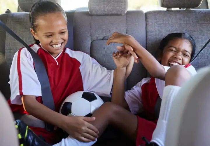 Two excited girls in soccer uniforms share a ball in the backseat, ready for their soccer fundraiser.