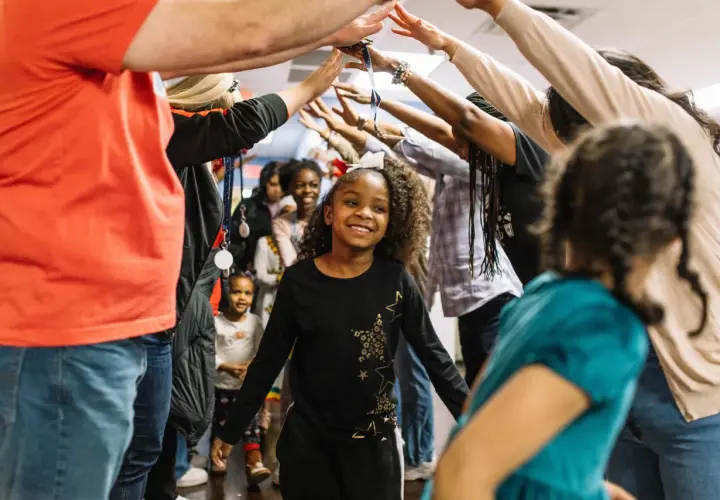 A group of children and adults form a tunnel with their arms raised, celebrating. This scene captures the joy of community and highlights places for kids to volunteer and make a difference.