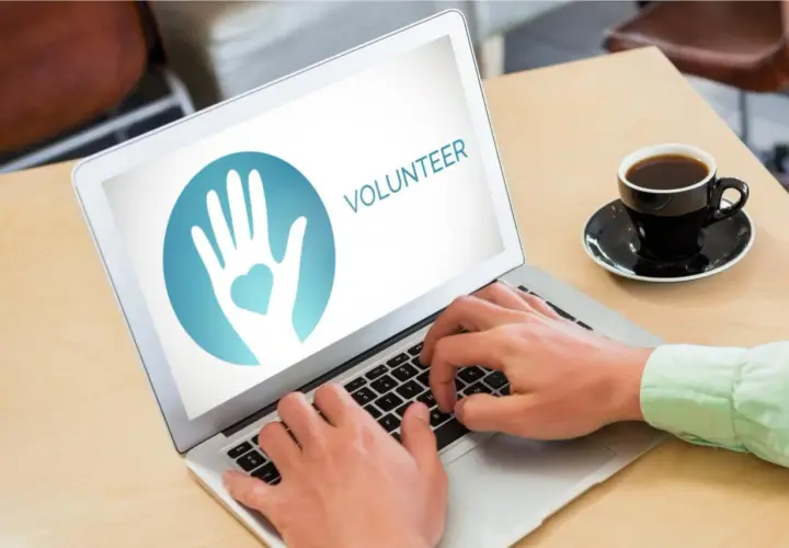 Hands typing on a laptop displaying a volunteer logo, ready to engage in online service projects. A coffee cup sits nearby.