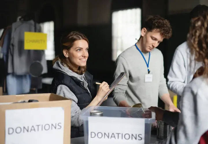 Volunteers organize donations for a free online community service. A woman smiles while checking items on a clipboard, while others sort canned goods.