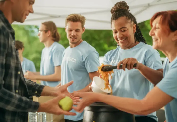 Volunteers serve food, showing what are some community services. A man receives an apple, while others get noodles.