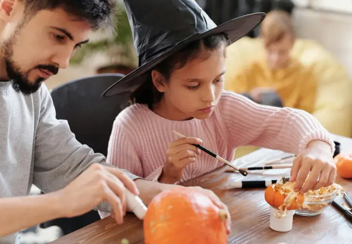 A girl in a witch hat and a man decorate pumpkins, showcasing fun Halloween fundraiser ideas.