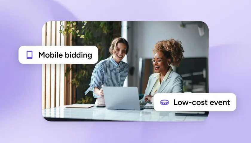 Two women smile while looking at a laptop, suggesting inexpensive silent auction ideas through mobile bidding for a low-cost event.