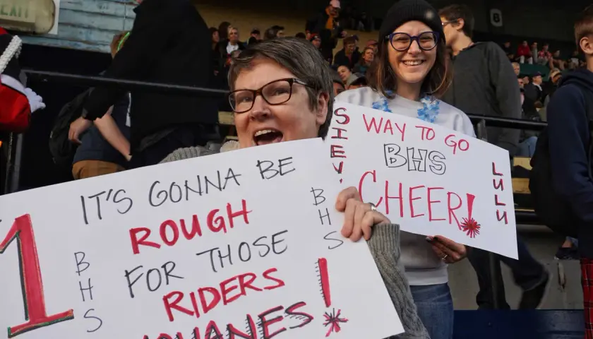 Fans hold signs at a snap raise fundraiser. One sign reads "IT'S GONNA BE ROUGH FOR THOSE RIDERS YOHANES!