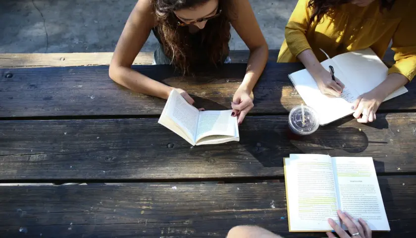 Two women study at a picnic table, one reading a book, the other writing notes. A board retreat activity.