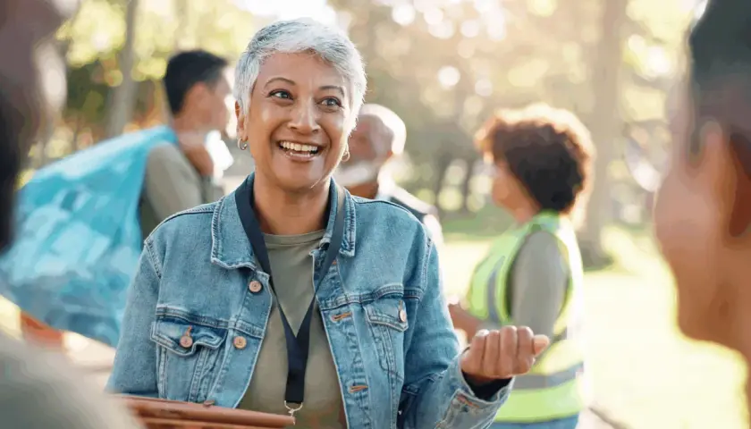 A woman with short gray hair smiles, her eyes bright. She's wearing a denim jacket and talking to others in a park, perhaps discussing why do you want to be a volunteer.