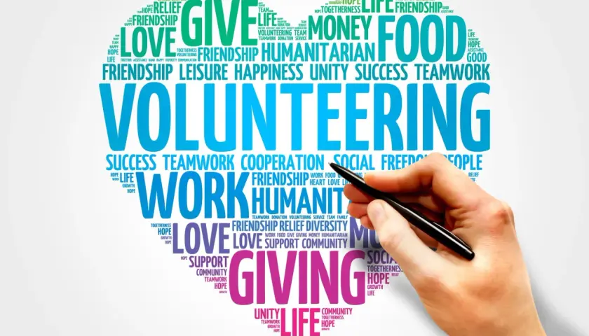 A hand holds a pen over a word cloud about volunteering, highlighting themes like "giving," "love," and "teamwork." This image represents the power of online volunteering.