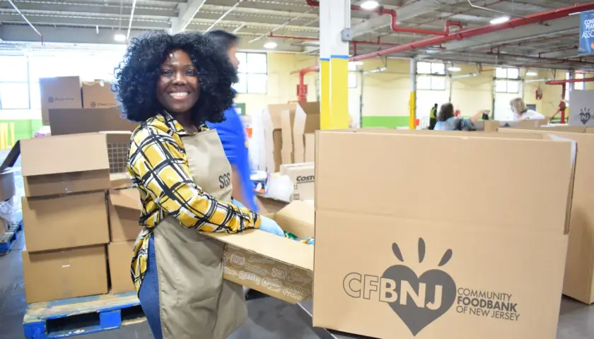 A smiling woman in an apron and gloves packs boxes at a food bank, embodying the spirit of a food bank volunteer job description.