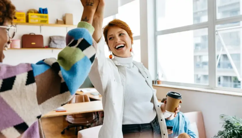 Two women high-five in an office, celebrating a successful project. One holds a coffee cup, perhaps after securing a moz nonprofit discount.