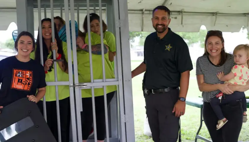 Smiles abound at a jail and bail fundraiser. A woman in a "Jail and Bail" shirt stands by a cage with women in neon shirts. A deputy and a woman holding a baby smile nearby.