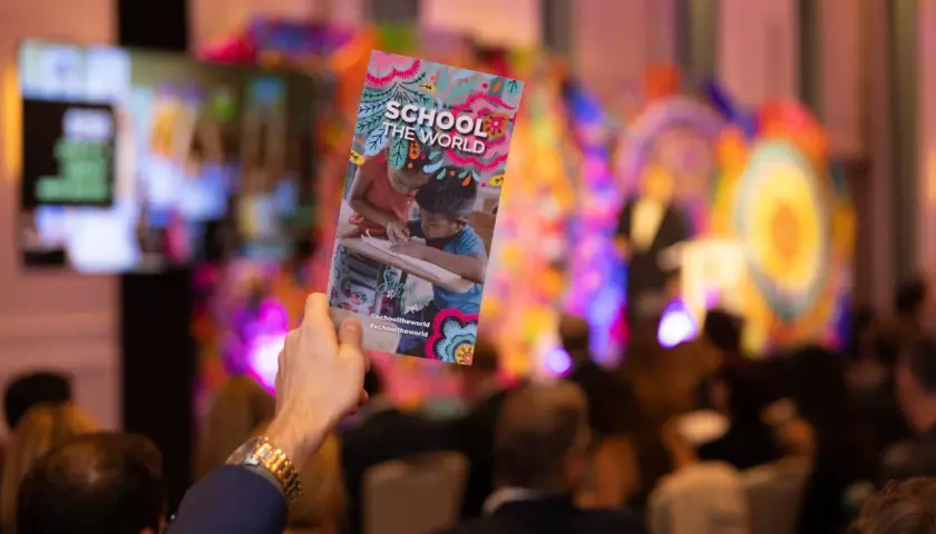 A hand holds up a colorful gala invitation for "School the World" at a vibrant event.