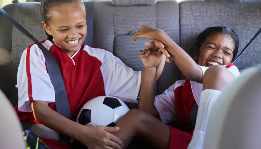 Two excited girls in soccer uniforms share a ball in the backseat, ready for their soccer fundraiser.
