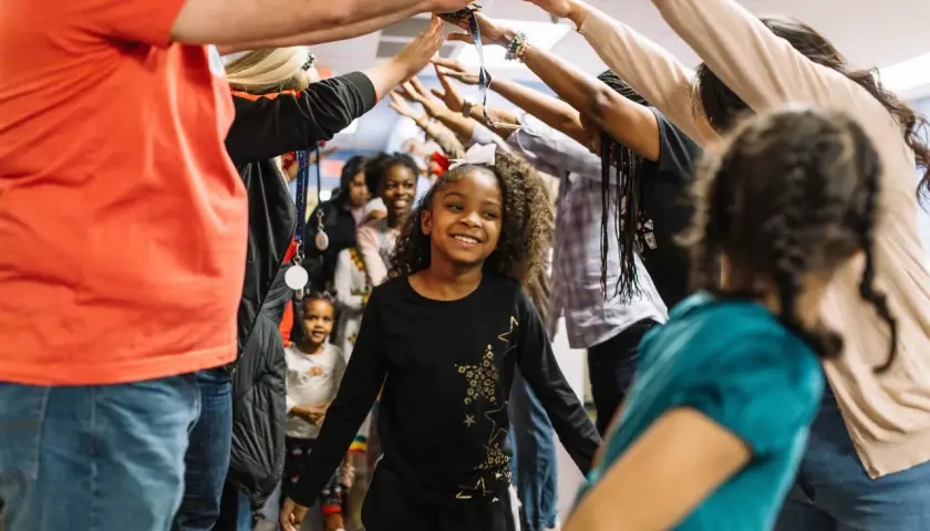 A group of children and adults form a tunnel with their arms raised, celebrating. This scene captures the joy of community and highlights places for kids to volunteer and make a difference.