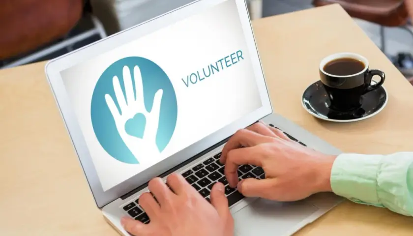 Hands typing on a laptop displaying a volunteer logo, ready to engage in online service projects. A coffee cup sits nearby.