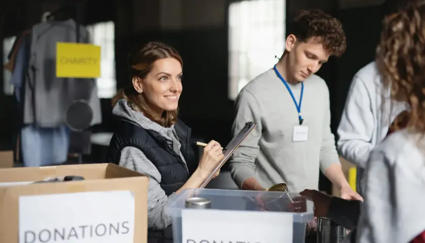 Volunteers organize donations for a free online community service. A woman smiles while checking items on a clipboard, while others sort canned goods.