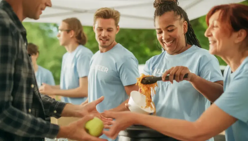 Volunteers serve food, showing what are some community services. A man receives an apple, while others get noodles.