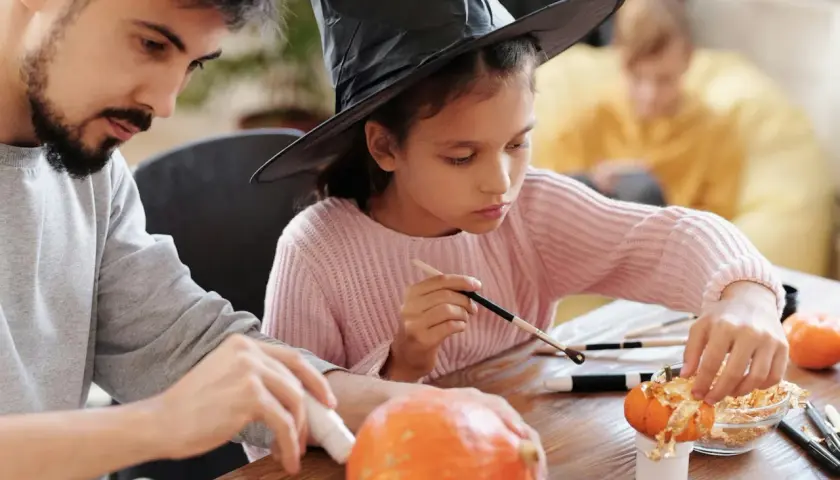 A girl in a witch hat and a man decorate pumpkins, showcasing fun Halloween fundraiser ideas.