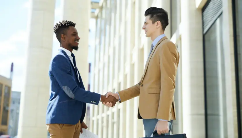 Two businessmen shake hands outside a modern building, perhaps after a successful meeting facilitated by their board portal.