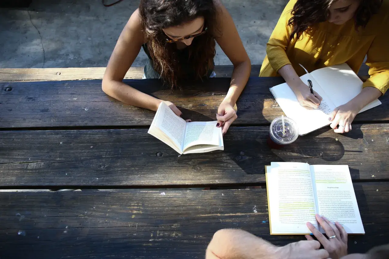Two women study at a picnic table, one reading a book, the other writing notes. A board retreat activity.