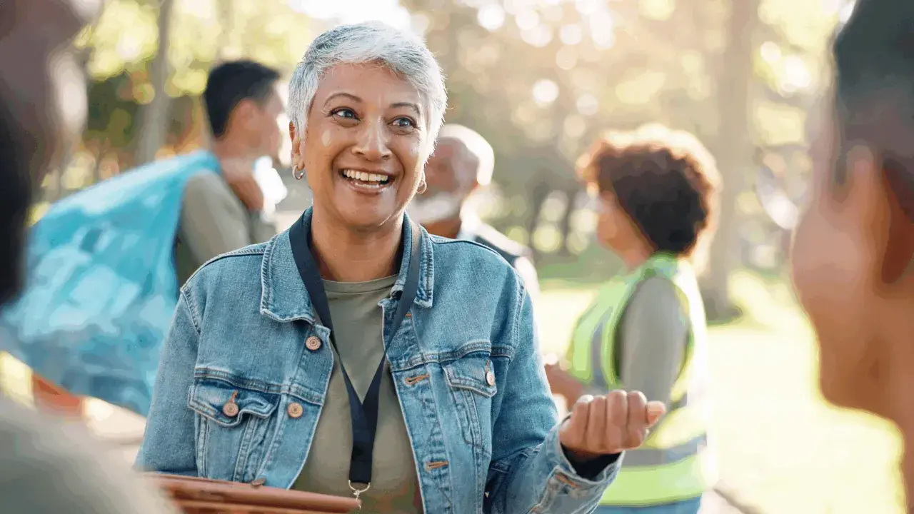 A woman with short gray hair smiles, her eyes bright. She's wearing a denim jacket and talking to others in a park, perhaps discussing why do you want to be a volunteer.