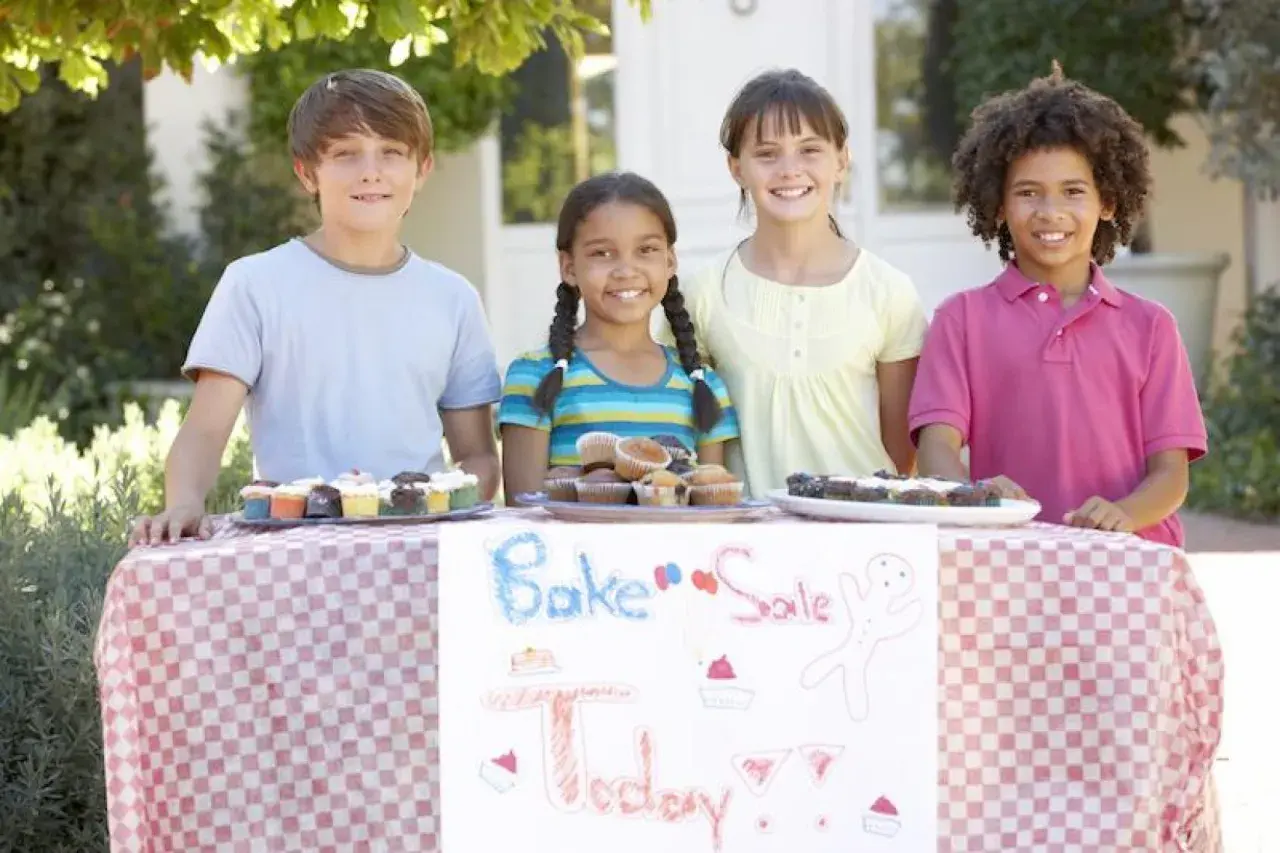 Four kids at a bake sale table, showcasing delicious cupcakes and brownies. Great fundraising ideas for 14 year olds!