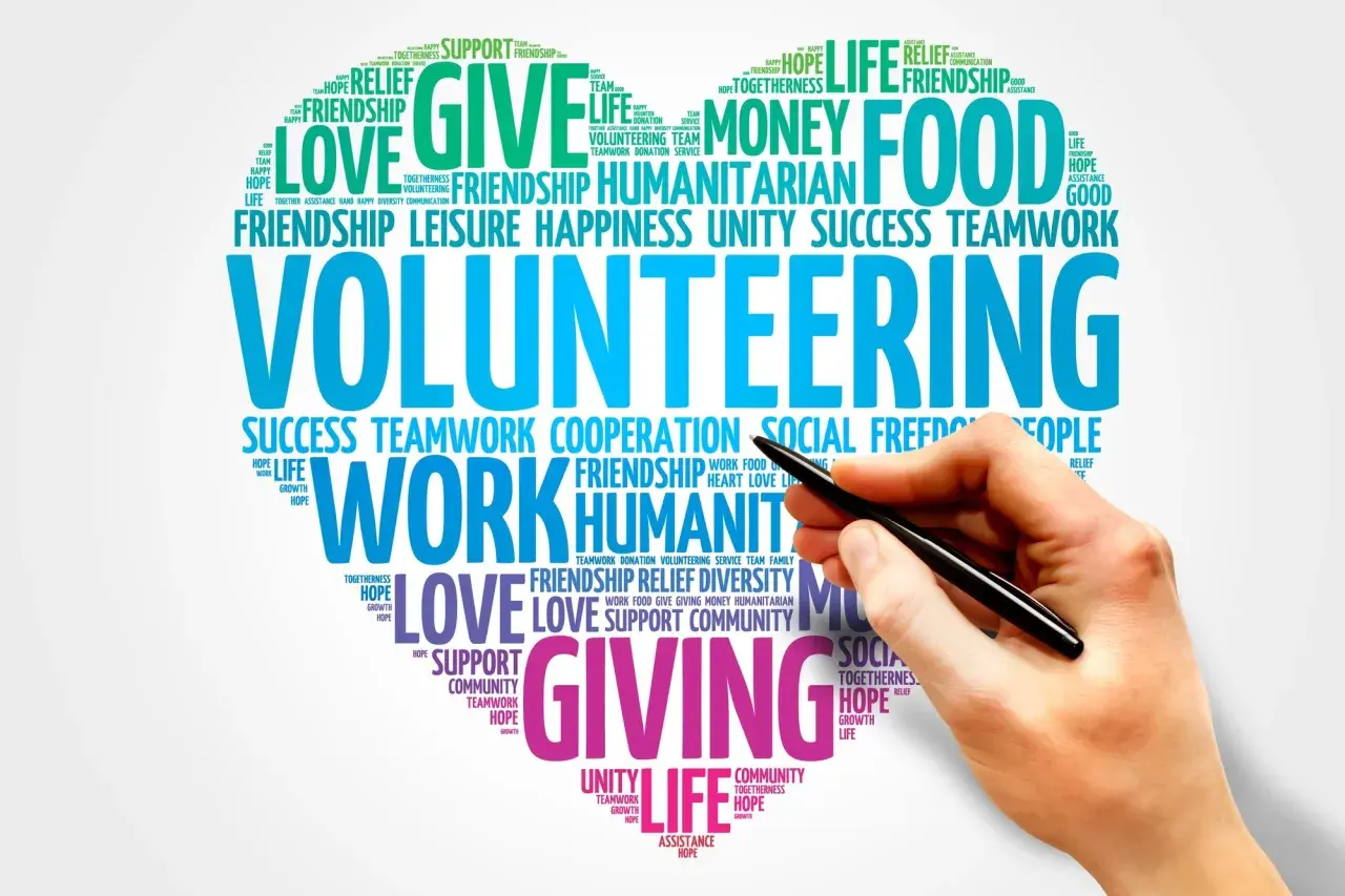 A hand holds a pen over a word cloud about volunteering, highlighting themes like "giving," "love," and "teamwork." This image represents the power of online volunteering.