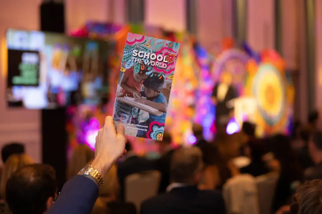 A hand holds up a colorful gala invitation for "School the World" at a vibrant event.