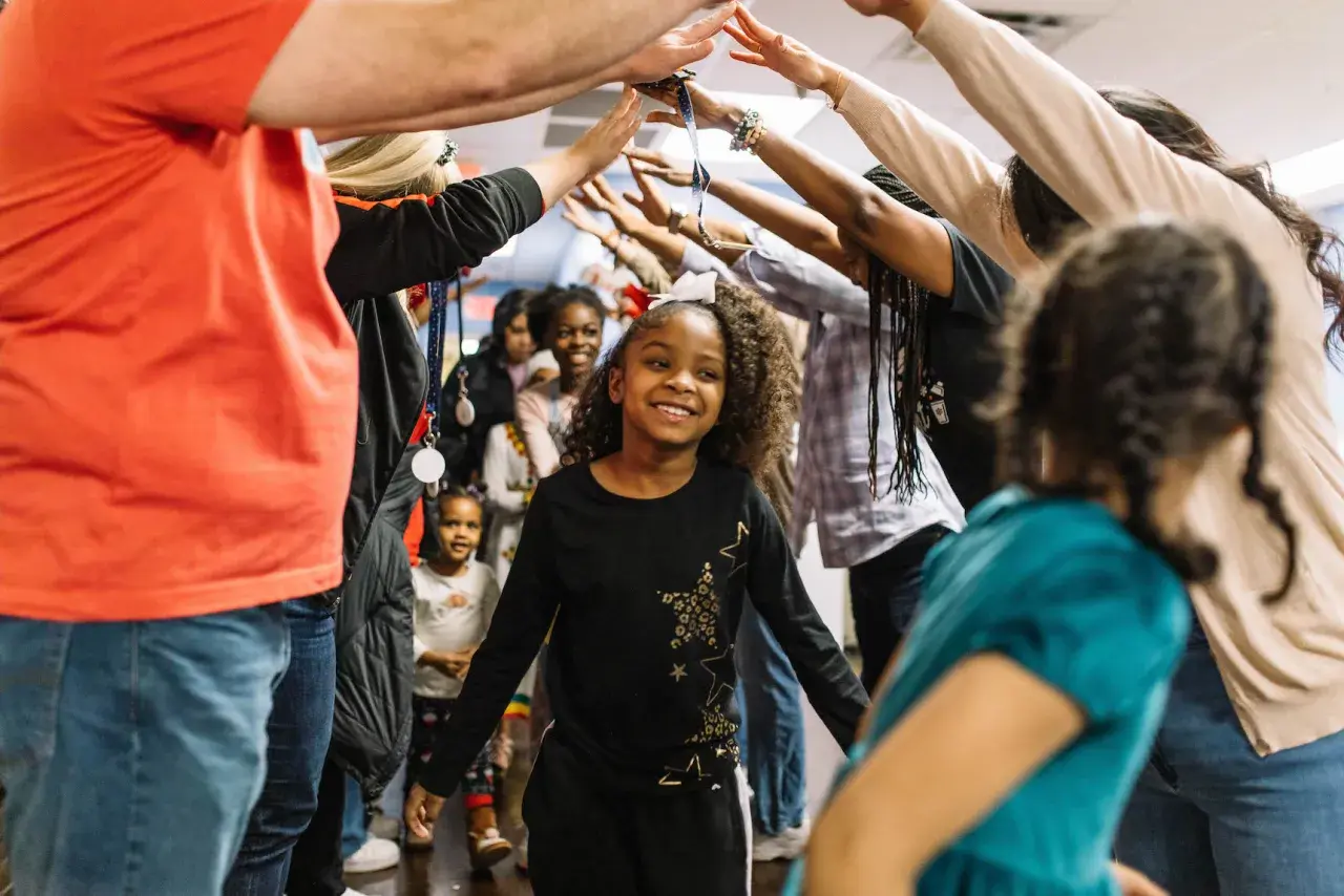 A group of children and adults form a tunnel with their arms raised, celebrating. This scene captures the joy of community and highlights places for kids to volunteer and make a difference.