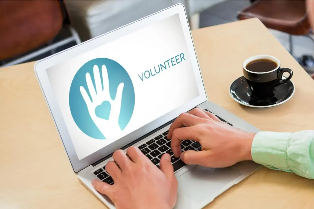 Hands typing on a laptop displaying a volunteer logo, ready to engage in online service projects. A coffee cup sits nearby.